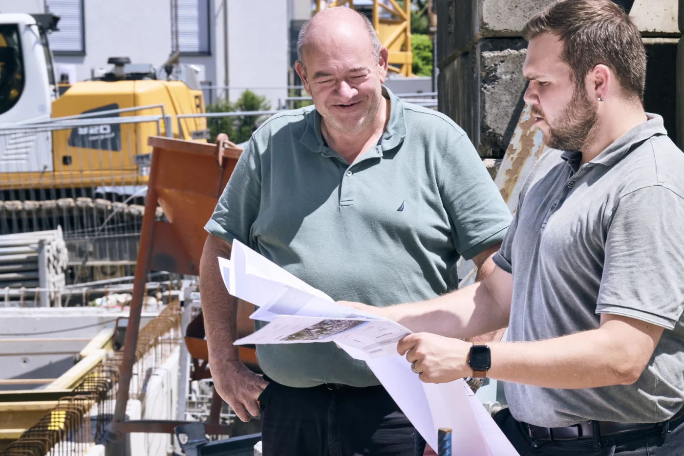 Zwei Männer in grauen Poloshirts stehen auf einer Baustelle, betrachten Baupläne und besprechen diese. Im Hintergrund sind Baumaschinen und Baumaterialien zu sehen.