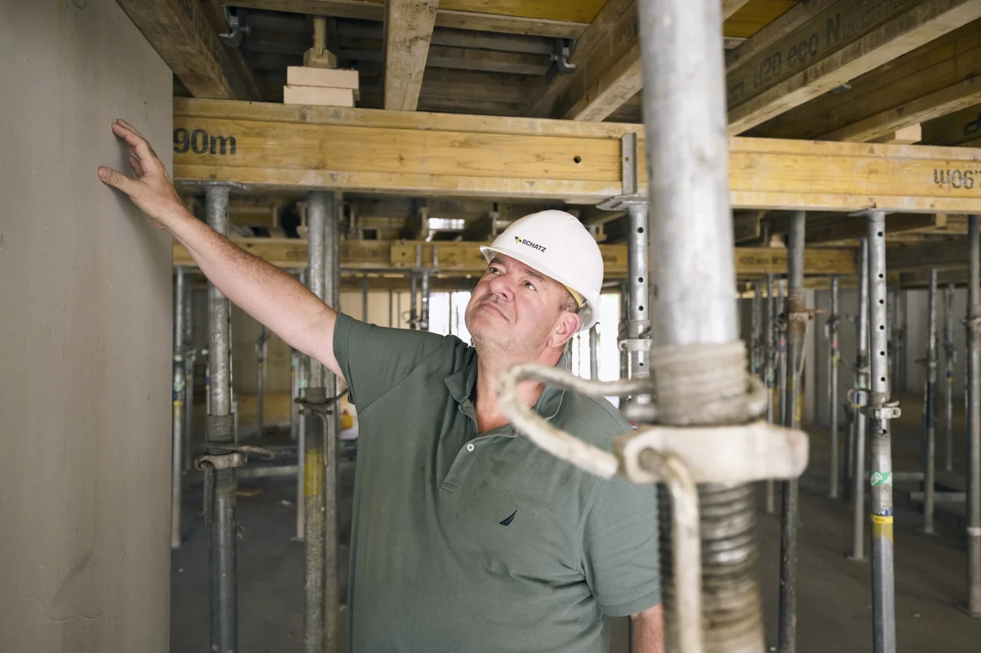 Ein Bauarbeiter in einem grünen Polohemd und einem weißen Schutzhelm begutachtet eine Wand im Inneren eines im Bau befindlichen Gebäudes, das von Metallstützen und Holzbalken umgeben ist.