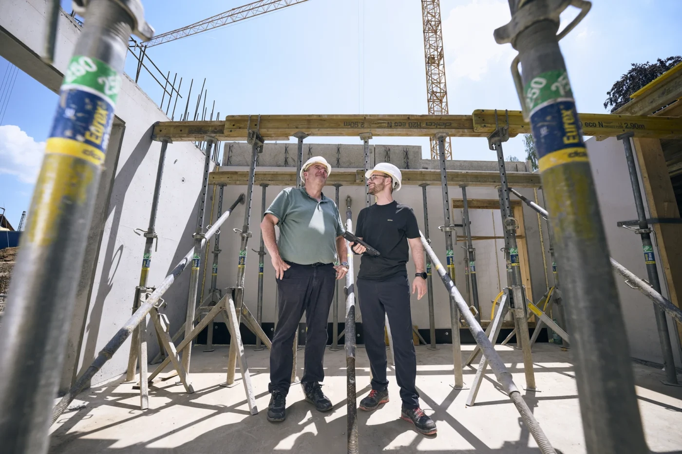 Zwei Männer mit Schutzhelmen und in Freizeitkleidung stehen auf einer Baustelle und unterhalten sich, umgeben von Metallgerüsten und unfertigen Betonwänden unter einem strahlend blauen Himmel.