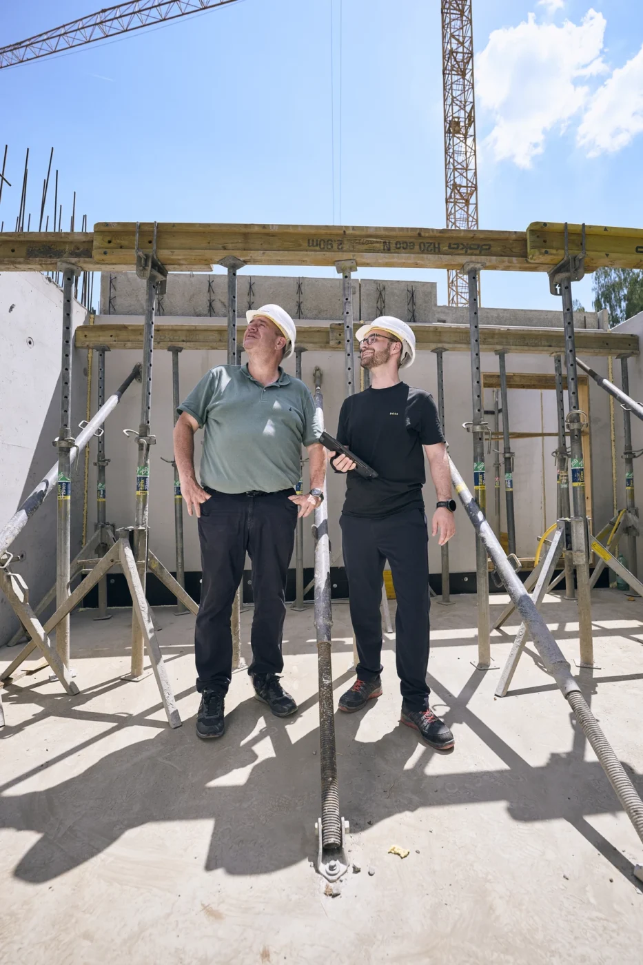 Zwei Männer mit Schutzhelmen und in Freizeitkleidung stehen auf einer Baustelle und blicken nach oben. Im Hintergrund sind unter einem sonnigen Himmel Metallgerüste, Betonwände und Kräne zu sehen.